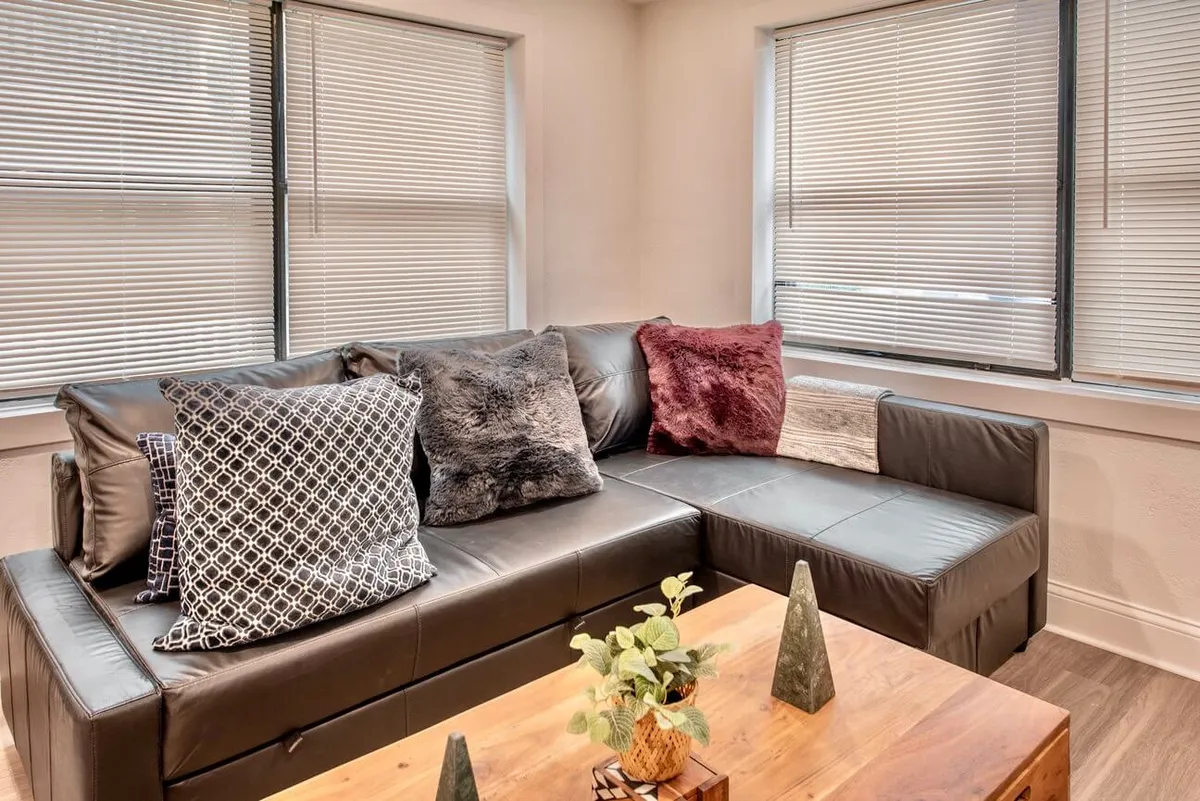 A stylish and comfortable living space featuring a black leather couch adorned with decorative pillows, a wooden coffee table, and small green plants. The room is arranged to feel inviting and suitable for social gatherings or quiet relaxation.