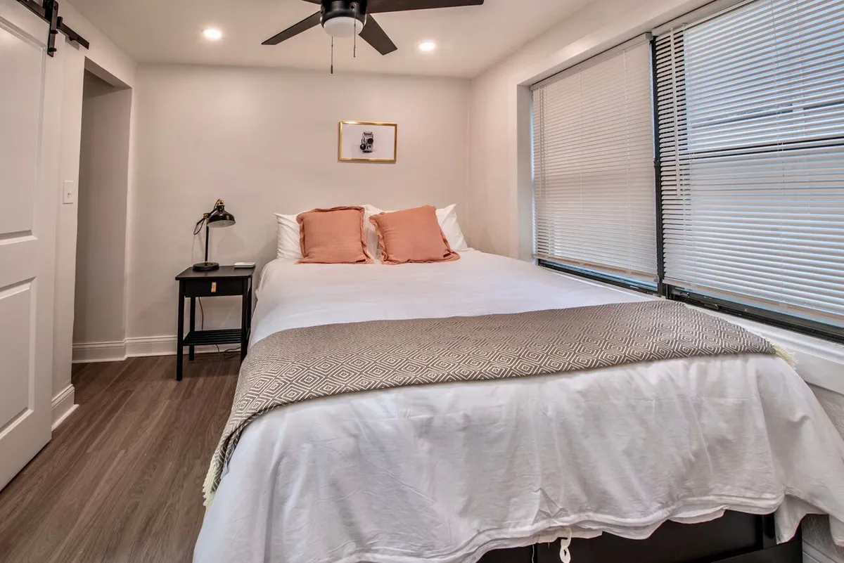 A quiet bedroom scene featuring a neatly made bed with plush pillows, a nightstand, subtle wall art, and soft morning light streaming through a window with blinds. The overall ambiance is peaceful and inviting, highlighting comfortable bedding and a soothing color palette.
